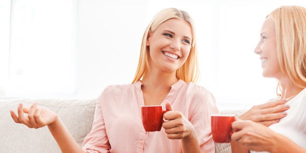 Happy young woman and her mother drinking coffee and talking while sitting on sofa together