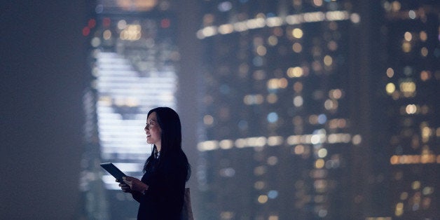 Joyful businesswoman looking away city skyline while using digital tablet, standing against illuminated highrise corporate buildings at night time.