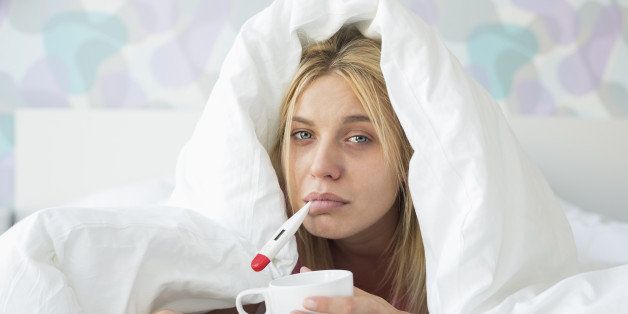 Portrait of sad woman with coffee mug taking temperature while wrapped in quilt on bed