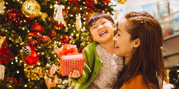 Pretty young mom carrying lovely little daughter who has a beautifully wrapped Christmas present in her hand while they talking to each other joyfully in front of a decorated Christmas tree in the shopping mall