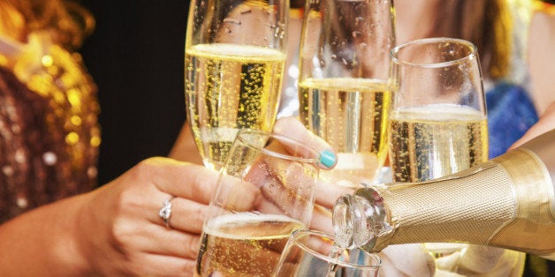 Five hands holding five champagne flutes being clinked together while being filled with champagne from a bottle. Glittery party clothing can be seen in the background being worn by the people holding the champagne flutes, but the faces of these people can not be seen. The lighting effect in the image is bright and sparkling.