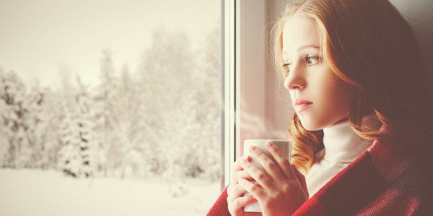 Pensive sad girl with a warming drink looking out the window in the winter forest
