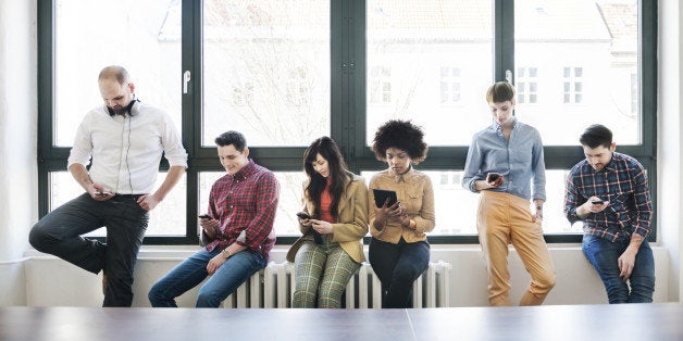 A group of young business people standing on a window talking and looking at their mobile devices. Backlit shot.