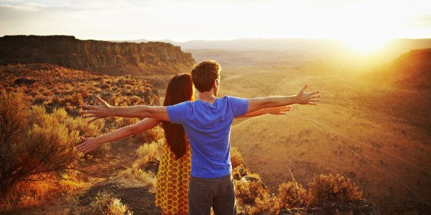 Couple standing on edge of desert canyon watching sunset with arms outstretched