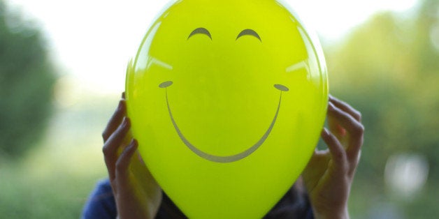 Portrait of a woman in a garden holding a green balloon in front of her face, with a happy face painted on it.