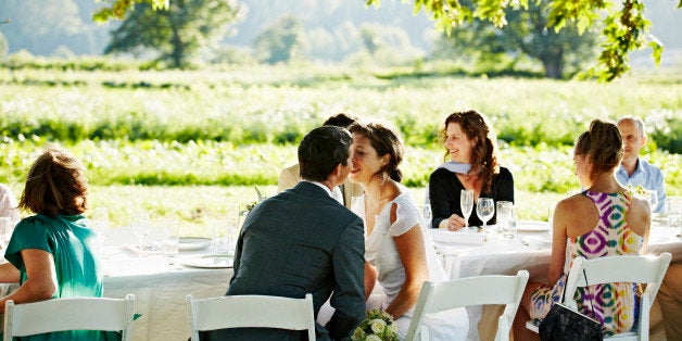 Bride and groom seated at outdoor banquet table about to kiss wedding guests in background