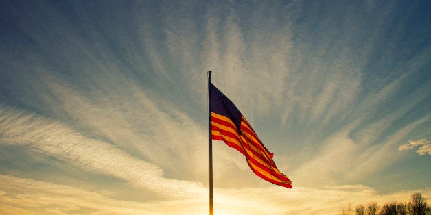 The sun sets behind a large flapping American flag under a blue sky.