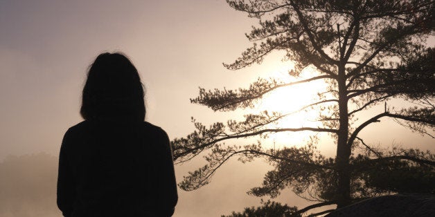 Silhouette of a woman sitting on rocks and facing sunrise sun, tranquil nature scenery, relaxation and meditation spiritual concept