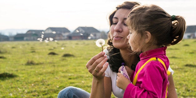 A woman with little girl dressed in casual clothing are blowing dandelion in the field with green grass. There are some distant village houses on the background of the image. The woman is dressed in white t-shirt and blue jeans, the girl is in a rose high neck long sleeve t-shirt with yellow suspenders and jeans.
