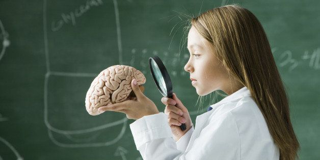 girl in a classroom standing in front of a chalkboard looking at a brain with a magnifying glass