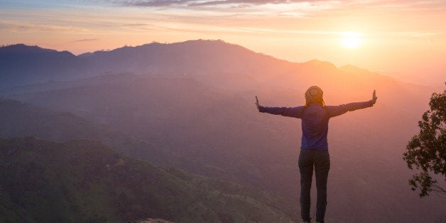 Happy celebrating winning success woman at sunset or sunrise standing elated with arms raised up above her head in celebration of having reached mountain top summit goal during hiking travel trek.