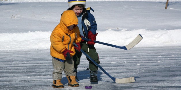 Little brothers have fun competing for a hockey puck on a outdoor pond. Each boy holds a small hockey stick and tries to hit a plastic purple puck. They wear winter jackets and boots. The picture was taken in Airdrie, Alberta, Canada. The background is snow covered.