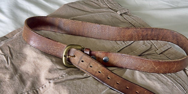 A man's belt shows the many marks of weight loss. Six adjustments / holes equates to six inches of weight loss. The belt sits on pants layer out on sheets of a bed, ready to be put on.