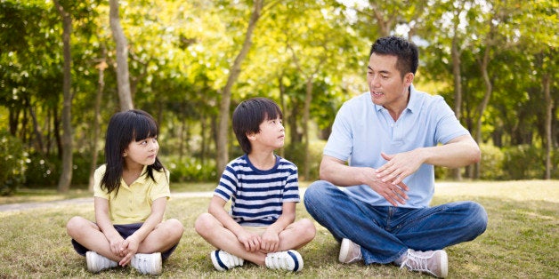 asian father and two children sitting on grass having an interesting conversation, outdoors in a park.