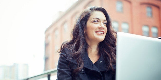 A happy young beautiful professional sits at an outdoor cafe in the city, holding a cup of espresso coffee and smiling while taking a break from her laptop computer. Horizontal with copy space. SELECTIVE FOCUS ON FACE.