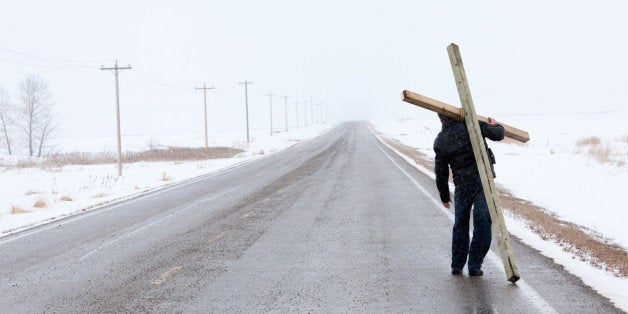 A man carrying a cross and walking down a desolate highway in winter. Easter theme. Additional themes are religion, spirituality, forgiveness, salvation, grace, mercy, love, crucifixion, burdens, suffering, journey, hope, and Good Friday. Unrecognizable Caucasian man in his 30s. This image is a remote highway in Alberta, Canada during a spring snow storm. Panorama.