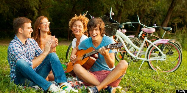 Young happy mixed race couples having romance fun outdoors at picnic. Young man play guitar to his woman. Flirting. Selective focus to smiling woman's face.