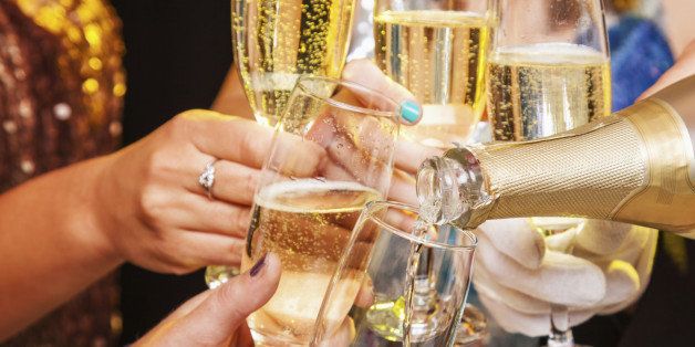 Five hands holding five champagne flutes being clinked together while being filled with champagne from a bottle. Glittery party clothing can be seen in the background being worn by the people holding the champagne flutes, but the faces of these people can not be seen. The lighting effect in the image is bright and sparkling.