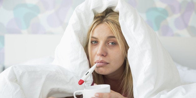 Portrait of sad woman with coffee mug taking temperature while wrapped in quilt on bed