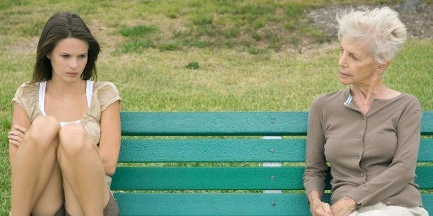 Teenage girl sitting apart from grandmother on bench, arms folded, looking down