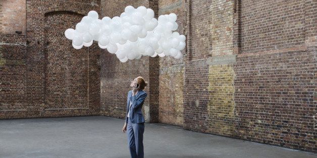 Businesswoman using mobile in empty warehouse with cloud made of balloons above head