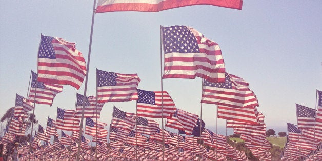 One main American flag, with a field of many American flags behind it, blowing in the wind on a sunny day with blue skies and sun flare.