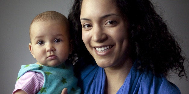 Adult woman with dark curly hair smiling and holding a baby with serious expression. Baby's eyes open looking directly into camera.