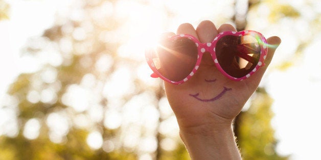 Hand with a smiling face drawn on palm, holding a pair of heart shaped sunglasses up to the sun.