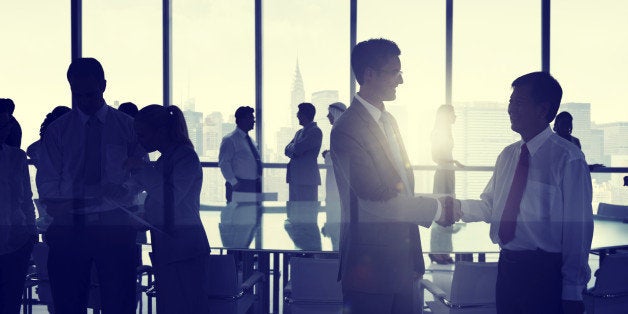 Business people shaking hands in an office building against a New York skyline. There is a large desk in the center of the room surrounded by office chairs.