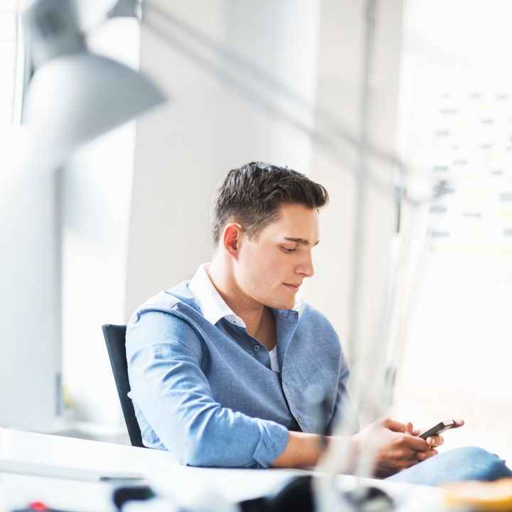 Young casual businessman sitting on his desk and texting on smart phone