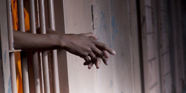 Cropped view of a prisoner in an orange jumpsuit standing in a prison cell with arms extended through the bars. Horizontal format.