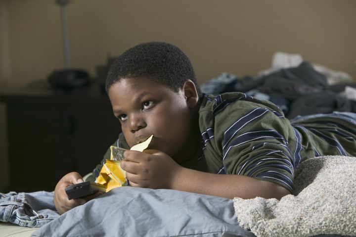 medium shot of a male child as he lays on his bed with the remote control and eats chips