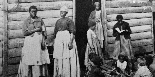 A group of women and children, presumably slaves, sit and stand around the doorway of a rough wooden cabin, Southern United States, mid 19th Century. One girl reads a book to the group of sitting children. (Photo by Hulton Archive/Getty Images)