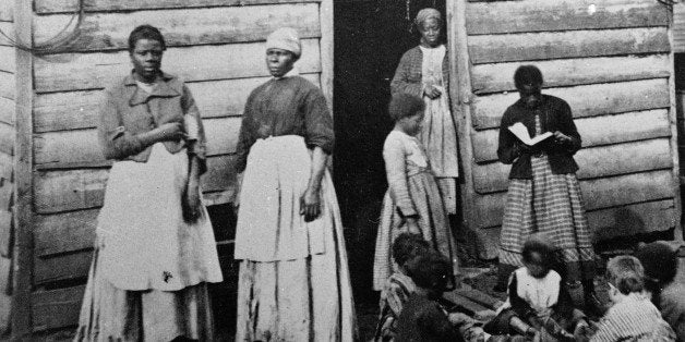 A group of women and children, presumably slaves, sit and stand around the doorway of a rough wooden cabin, Southern United States, mid 19th Century. One girl reads a book to the group of sitting children. (Photo by Hulton Archive/Getty Images)