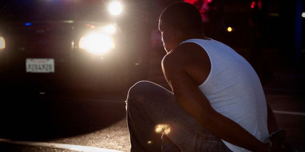 Police shining lights on handcuffed African man sitting on curb