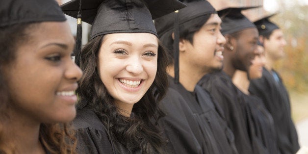 Six multi-ethnic friend graduates excitedly wait for their name to be called during graduation ceremony. Mixed-race girl looks back at camera. School building background.