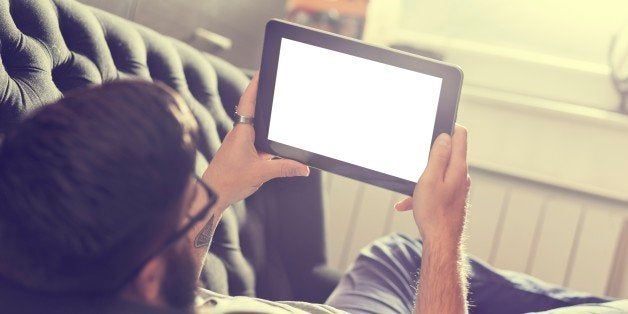 Male model lying on a couch in a living room, surfing the web on a tablet computer