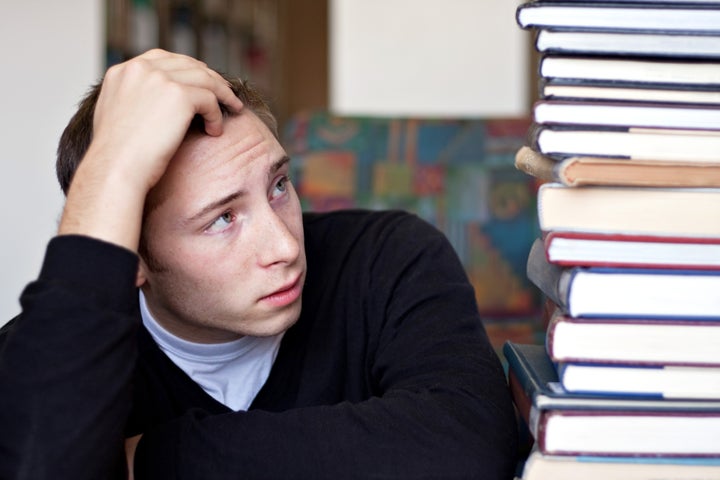 A frustrated and stressed out student looks up at the high pile of textbooks he has to go through to do his homework.