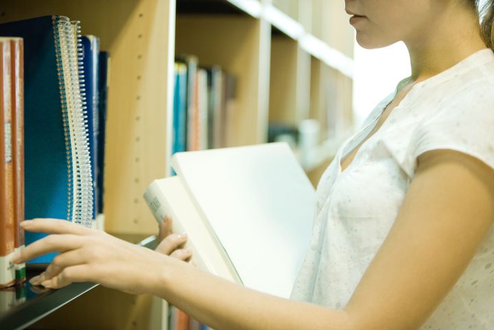 Young woman reaching for books on shelf in library, cropped view