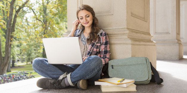 A female college student sits outside a campus building with her books and a laptop computer. She is placing earbuds in her ears.