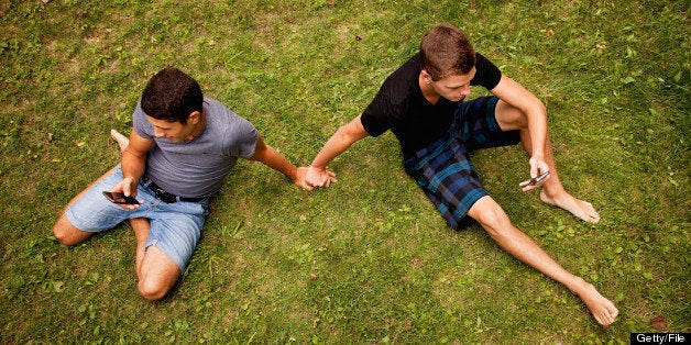 Teen boys sitting on the grass using their cellphones, holding hands.