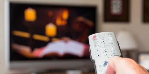 Silver modern TV remote control being pressed by thumb with out of focus screen background