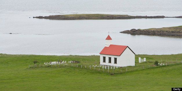 Small, white church at the edge of a body of water, surrounded by green grass, Iceland.