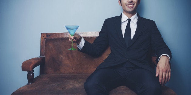 Smiling young businessman is sitting on a vintage style sofa with a martini glass in his hand