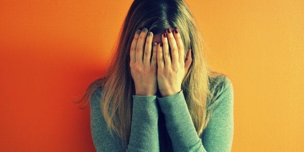 Woman closing her face with her hands, she has different red and blue nailpolish, standing against bright orange wall