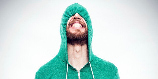 A bearded hipster young man pulls the drawstrings on his green hoodie, partially concealing the funny face he is making, comp