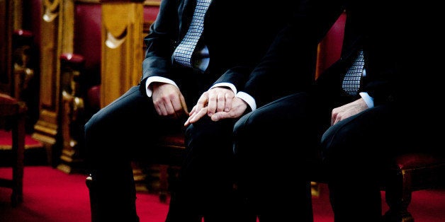 Civil gay marriage, 2 men holding hands while sitting on the chairs, wearing black suits. Background is brown and red, horizo