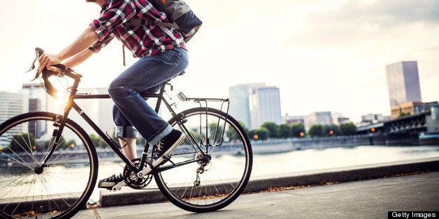 A young man commuting in an urban city environment on his street bicycle, a waterproof bike bag on his back. This is the Eastbank Esplanade in Portland, Oregon, that follows along the Willamette river. Downtown is visible across the water, the sun shining brightly between the buildings. Horizontal, in-camera lens flare, in motion.