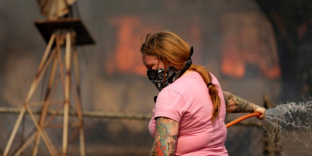 A woman douses water from a hose around her home as her neighbor's home burns during a wildfire Thursday, May 15, 2014, in Escondido, Calif. One of the nine fires burning in San Diego County suddenly flared Thursday afternoon and burned close to homes, trigging thousands of new evacuation orders.(AP Photo/Gregory Bull)