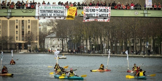 Indigenous people from South America paddle on kayaks as others stand on a bridge holding banners reading 'Native territories